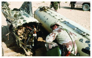 A soldier in desert camouflage inspects the wreckage of a Scud missile that has been destroyed. The missile is broken in half on the sand, exposing its internal wiring and components.